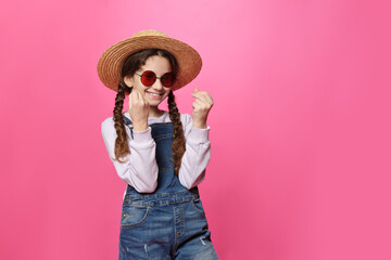 Cute little girl in hat and red sunglasses, looking at camera doing money gesture with hands, isolated pink background.