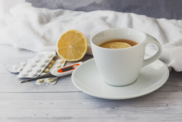 White cup with tea with lemon,  thermometer and pills in a blister on wooden background. Seasonal winter, autumn illnesses, colds or flu. Home alternative treatment concept.