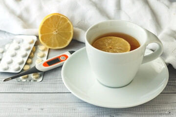 White cup with tea with lemon,  thermometer and pills in a blister on wooden background. Seasonal winter, autumn illnesses, colds or flu. Home alternative treatment concept.