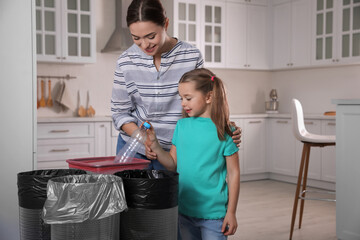 Young woman and her daughter throwing plastic bottle into trash bin in kitchen. Separate waste...
