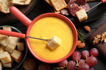 Pot of tasty cheese fondue and snacks on black wooden table, flat lay