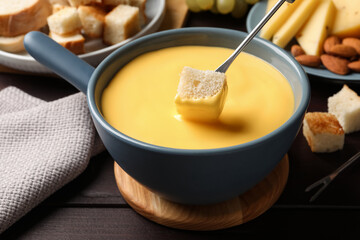 Dipping piece of bread into tasty cheese fondue at wooden table, closeup