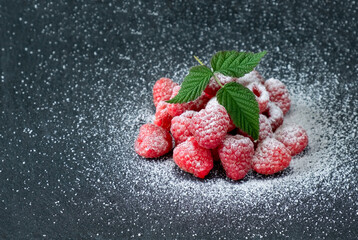Ripe fresh raspberries with sugar powder and green leaves on a gray stone table. Close-up view