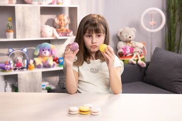 Obraz na płótnie Canvas Young brunette kid girl can not make right decision and choice what macaron to bite first - right from with lemon and beryy filling or left made with violet cream, then bite macaron in yellow color