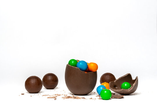 Cracked chocolate easter egg with colorful small round candies and chocolates on white background, copy space. Chocolate treat for kids. Easter concept.