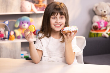 Lovely joyful school girl can not choose between a white donut and yellow fresh macaroon. Funny teenager girl having fun with donut and macaron at modern home. Sweet tooth. Unhealthy food concept