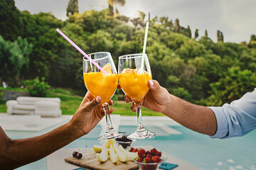 Hands clinking orange cocktail glasses against a swimming pool background - healthy aperitif in...