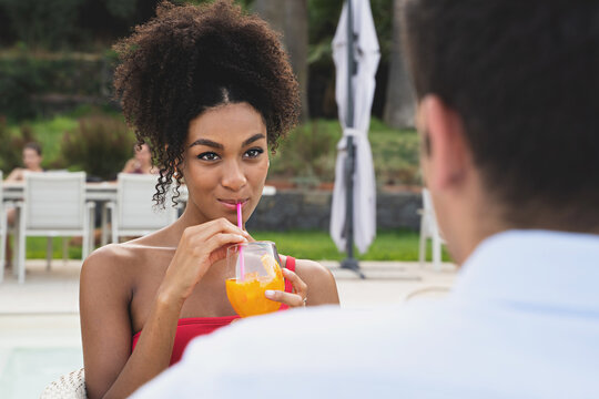 Romantic couple of multiethnic young people sitting by the swimming pool drinking juicy orange fruit smoothies - summer and vacation lifestyle concept