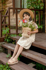 Dark-haired preteen girl sitting on doorstep of rural house with basket of wildflowers