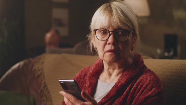 Shocked elderly female in glasses looking around after reading terrifying news on mobile phone while sitting on sofa in living room at home