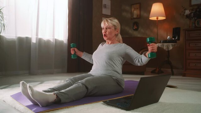 Aged female in sportswear watching tutorial video on laptop and exercising with dumbbells while sitting on floor during fitness workout in sunny morning