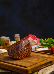 Appetizing ham in the foreground on a wooden board, in the background - raw meat, herbs, vegetables, spices. Dark blue background. Festive dish. Restaurant, banquet, recipes.