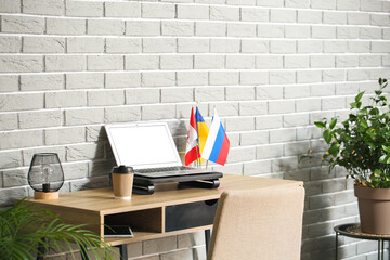 Laptop and flags of Russia, Ukraine and Canada on table near grey brick wall