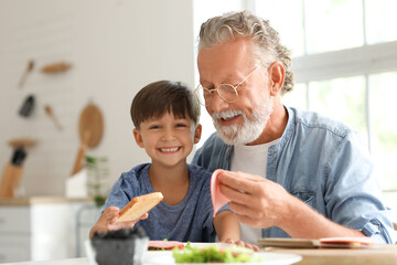 Little boy making sandwich with his grandfather at table in kitchen