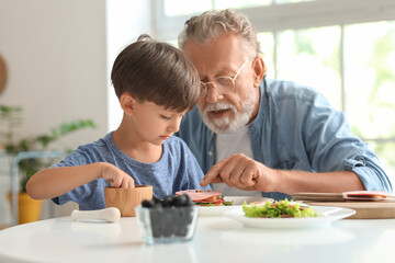 Little boy making sandwich with his grandfather at table in kitchen