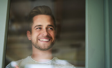 Make everyday an awesome one. Shot of a handsome young man looking out of a window at home and smiling.