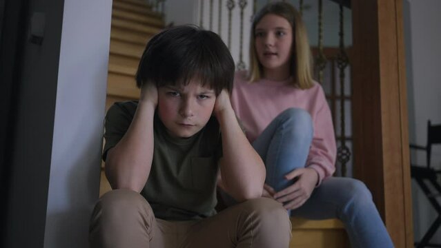 Dissatisfied Caucasian boy closing ears as displeased girl yelling sitting on stairs at background. Portrait of sad brother looking at camera as furious sister scolding sibling indoors at home