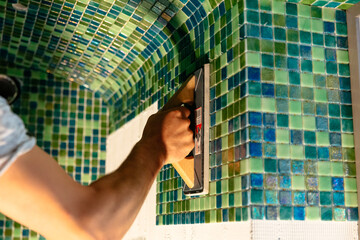 Worker applying mosaic tiles in bathroom, sauna walls. Construction tools and equipment. Spa or sauna relaxation modern room interior design. Adhesive for mosaic tile. Renovation concept - obrazy, fototapety, plakaty