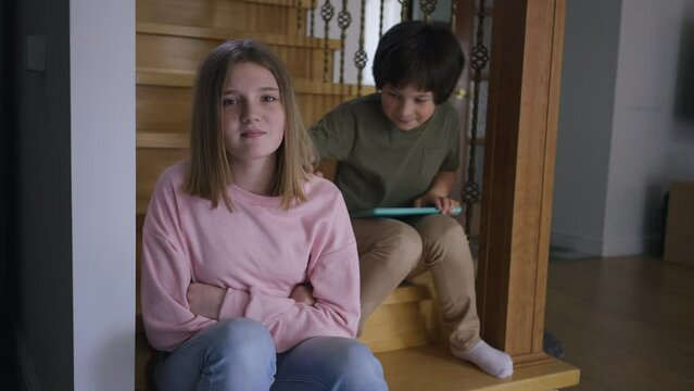 Portrait of tired teenage girl looking at camera with sad facial expression as little boy pushing shoulder. Upset Caucasian sister with annoying brother at home in living room. Family conflicts