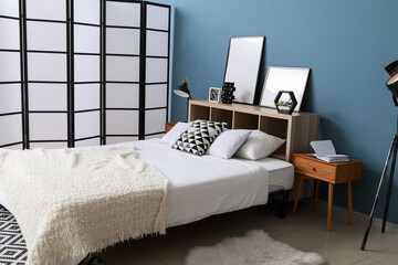 Comfortable bed, folding screen and blank posters on shelf near blue wall