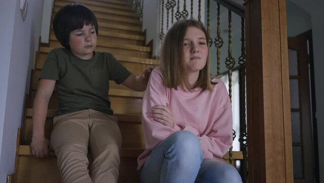 Irritated teenage girl pushing away hand of little boy leaving as sibling sitting on stairs hugging knees. Portrait of argued Caucasian sister and brother at home indoors. Rivalry and lifestyle