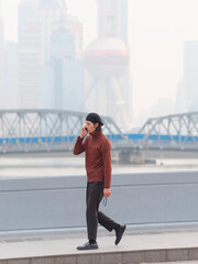 Portrait of handsome Chinese young man with sunglasses in hand walking and talking in mobile phone with Shanghai city landmarks background, male fashion, cool Asian young man lifestyle.