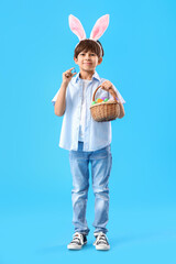 Cute little boy with bunny ears and Easter eggs in basket on color background