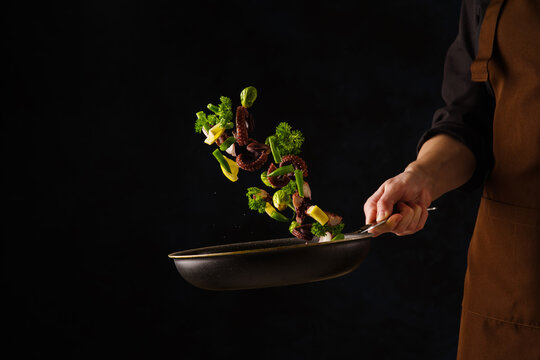 Seafood - Octopus With Vegetables And Herbs In A Pan In A Frozen Flight On A Black Background. Professional Chef Prepares Seafood. Restaurant Cuisine Recipes. Asian Seafood Cuisine.