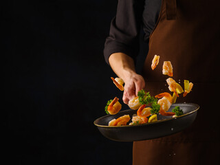 The chef in a dark uniform cooks seafood - shrimp with vegetables in a pan. Seafood in frozen flight on a black background. There is free space to insert. Seafood recipes. Restaurant, hotel, banner.