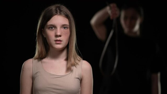 Frightened beaten teenage girl looking with fear back at blurred woman holding belt. Portrait of Caucasian bruised teenager posing at black background looking at camera. Domestic violence and rage