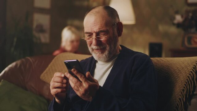 Pan around view of cheerful elderly male in glasses chuckling and reading news while sitting on sofa, and browsing social media on smartphone on weekend day at home