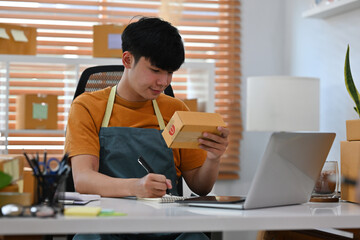 A portrait of an Asian man, e-commerce employee sitting in the office full of packages in the background looking at the package and writing a note, for SME business, e-commerce and delivery business.
