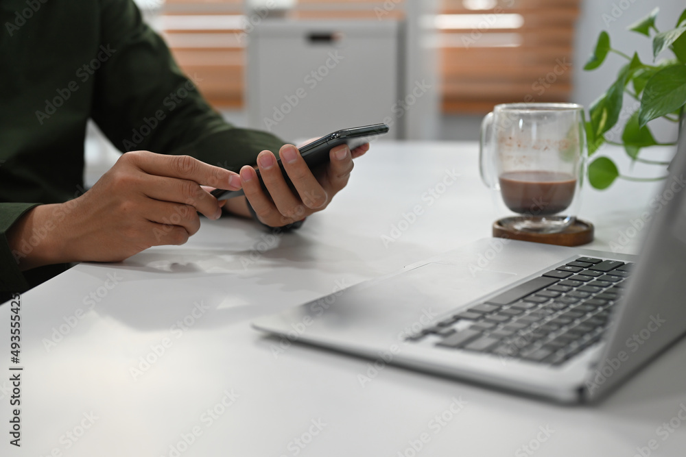 Poster side view close up of a young man sitting in the office typing on a smartphone and working on a lapt