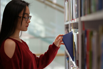 Asian student girl standing between library bookcases, young korean woman picking up book from shelf while studying in university library, using learning materials. Educational opportunities