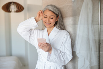 Pretty Asian woman in bathrobe halding smartphone in shower - obrazy, fototapety, plakaty