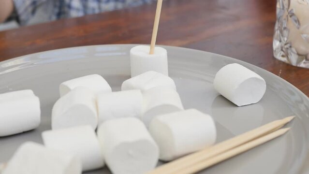 Close-up, DOF: teenagers stick marshmallows on sharp wooden sticks before roasting and frying on a burning candle. Slow motion.