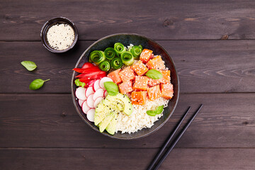 Poke bowl with salmon or tuna, rice, avocado, cucumber, radish, pepper, sesame seeds on dark wooden background. Hawaiian diet food with fish. Top view. Flat lay. 