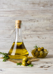 Golden olive oil in a clear jug on a canvas napkin. Green olives in a glass vase in the background. Sprigs of rosemary on the table