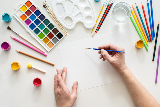 The female hands while drawing. View from above. artist's desk with colorful paints and pencils. woman draws with blue paint on a white sheet of paper. Drawing lesson Creative hobby of adults. 
