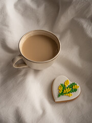 Morning coffee with milk with heart-shaped cookies painted with mimosa pattern icing  on a white bed in the morning light