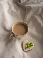 Morning coffee with milk with heart-shaped cookies painted with mimosa pattern icing  on a white bed in the morning light
