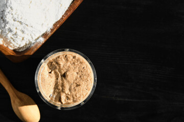 Active rye and wheat sourdough starter in glass jar with ingredients, flour . Top view.