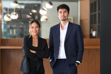 Two successful businesspeople in formal wear standing in modern office and looking confidently at camera.