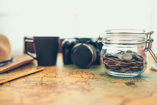 Travel money savings in a glass jar, camera, coffee cup, straw hat and pen on world map. Travel budget concept.