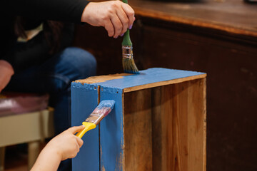 Closeup of young girl and woman hands with brushes slowly painting very old wooden closet drawer in...
