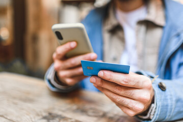Soft focus of crop man with cellphone using credit card to pay for purchase while sitting at table and shopping online in restaurant