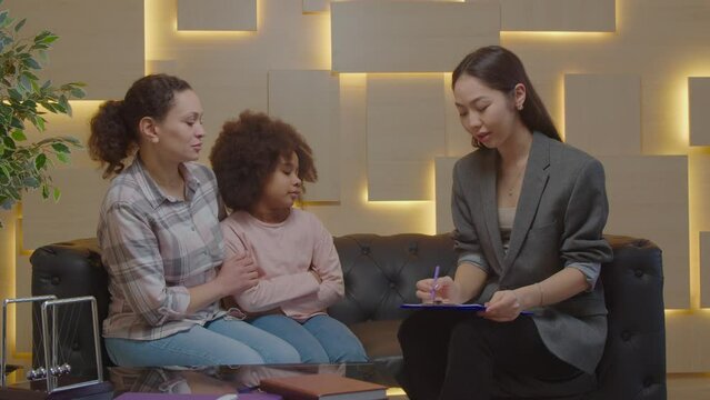 Worried affectionate middle eastern mother and adorable school age multiracial daughter with adolescent problems counselling with attentive pretty Asian psychologist, to improve social skills indoors.