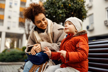 Little talkative boy sitting with his nanny of caregiver on the bench outside and telling a story.