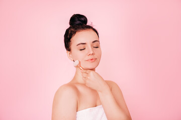 A relaxed young woman standing isolated on a pink background and smiling with her eyes closed, using a rose quartz facial roller. the concept of home care