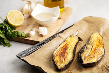 Ingredients for a traditional dish of arabic cuisine baba ganoush: roasted eggplant, tahin, cilantro, garlic, olive oil, lemon juice on wooden board and baking tray